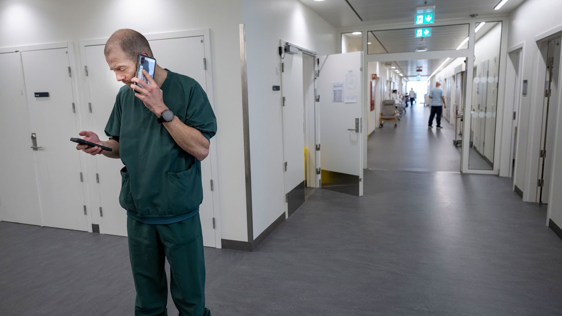 På operationsgangen i Hospitalsbyen. Jens Jakob Riis taler i telefon med cheflæge Helga Gulisano. De drøfter den behandling, han skal give en patient med aneurisme. På den anden telefon kan Jens Jakob Riis se opskriften på alteplase, som han skal blande til patienten. Foto: Lars Horn / Baghuset. 