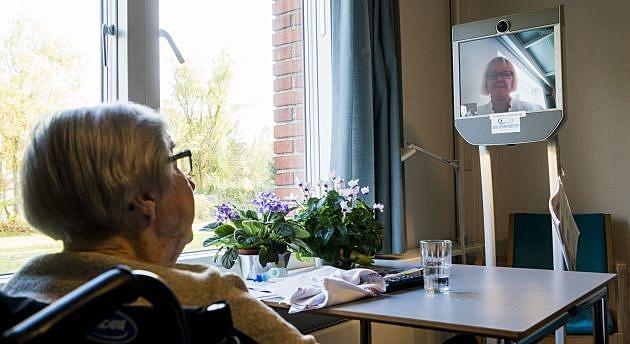 Grethe Pedersen på 92 år sidder i en stol ved vinduet på en af Aarhus Kommunes 22 korttidspladser. Med udsigt til løvfald og overlæge Catherine Foss på den rullende fladskærm. Foto: Carsten Ingemann