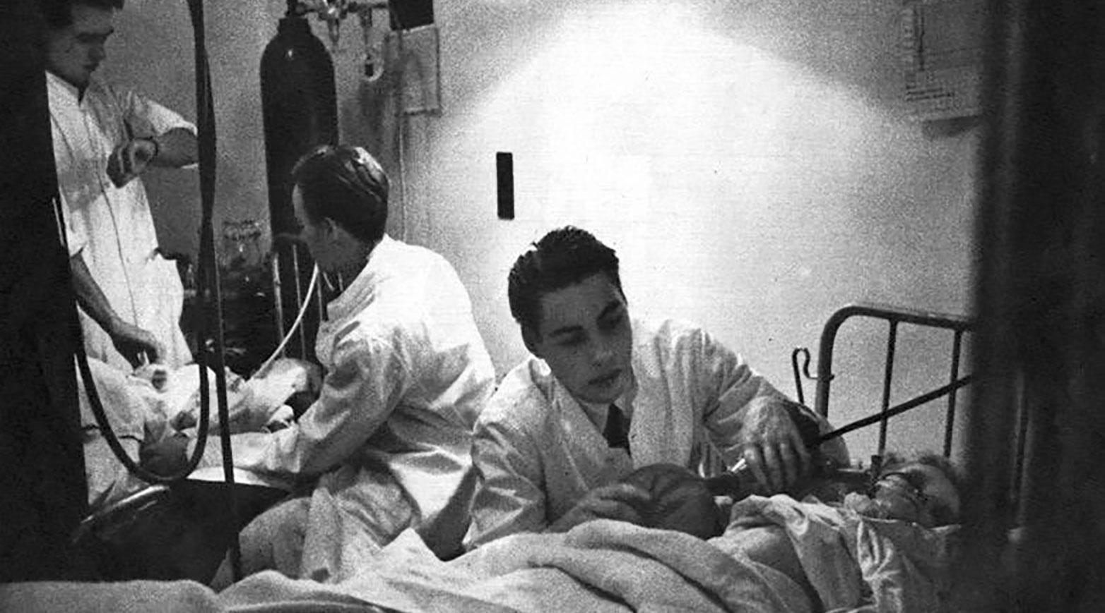 Medicinstuderende som frivillige ventilatører på Blegdamshospitalet under polioepidemien i 1952. Stående t.v. er Hans Erik Jensen ved at tage pulsen på en patient, mens Carl Eli Olsen (med ryggen til) passer ilttilførslen. I forgrunden har Svend M. Hanen blottet mellemgulvet på et barn for at tjekke det åndedræt, han selv skaber. Foto udlånt af Carl Eli Olsen.