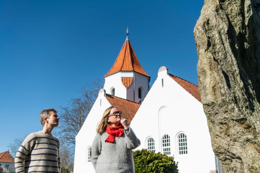 Line Modin og Mette Geil foran den dannebrogsfarvede frimenighedskirke ved mindestenen for de 16 unge mænd fra menigheden, der faldt i 1. Verdenskrigs skyttegrave i blandt andet Rusland og Frankrig. Foto: Palle Peter Skov 