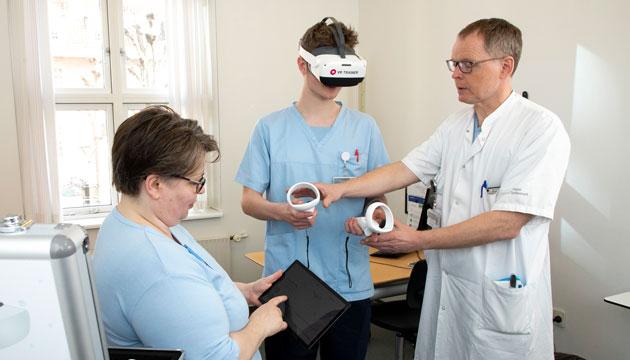Implementeringsleder Trine Ladingkær og overlæge Jens Kjeldsen instruerer en kollega i det VR-udstyr, der blandt andet bruges til at undervise i, hvordan man overholder hygiejnereglerne på isolationsstuer. Foto: Heidi Lundsgaard