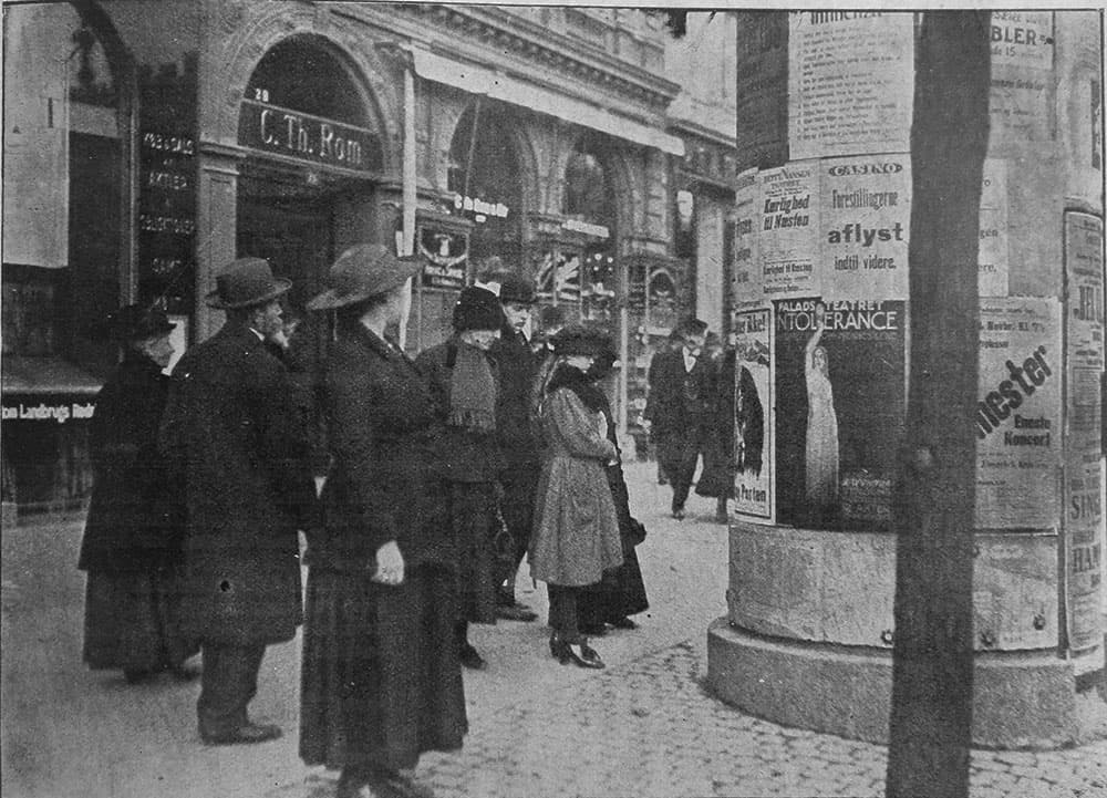 Københavnere betragter plakatsøjle med meddelelser om aflyste arrangementer og Sundhedsstyrelsens  12 hygiejneråd til befolkningen mod influenzasmitte (foto i Hver 8. Dag, den 1/11 1918).