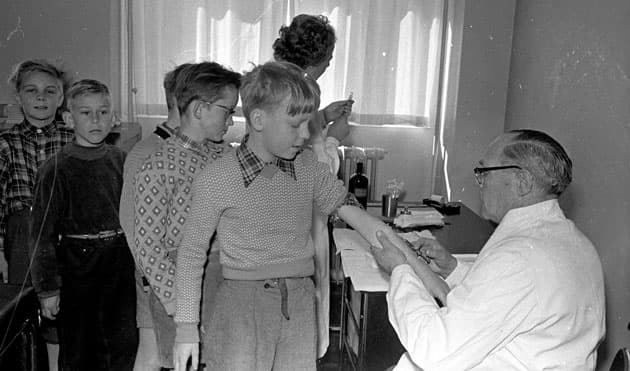 Efter polioepidemien i 1953 var danske forældre panisk angste for, at deres børn skulle blive ofre for en ny epidemi. Her er det amtslæge Hj. Heerup, som i april 1955 stikker nålen i armen på en betænkelig elev på Munkebjergskolen i Odense, mens kammeraterne bagved nervøst venter på deres tur. Foto: Fyens Stiftstidendes pressefotosamling, Odense Stadsarkiv