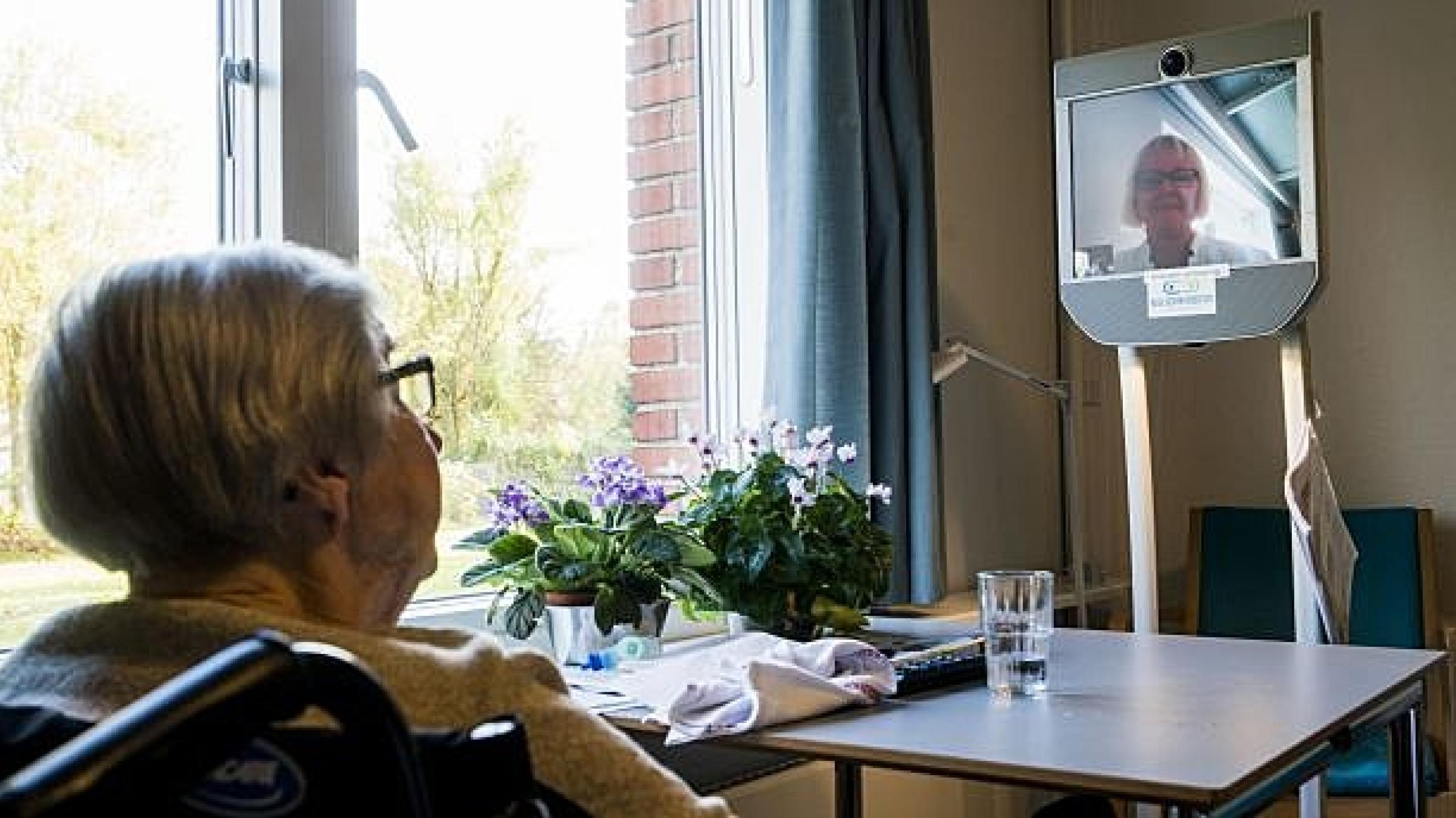 Grethe Pedersen på 92 år sidder i en stol ved vinduet på en af Aarhus Kommunes 22 korttidspladser. Med udsigt til løvfald og overlæge Catherine Foss på den rullende fladskærm. Foto: Carsten Ingemann