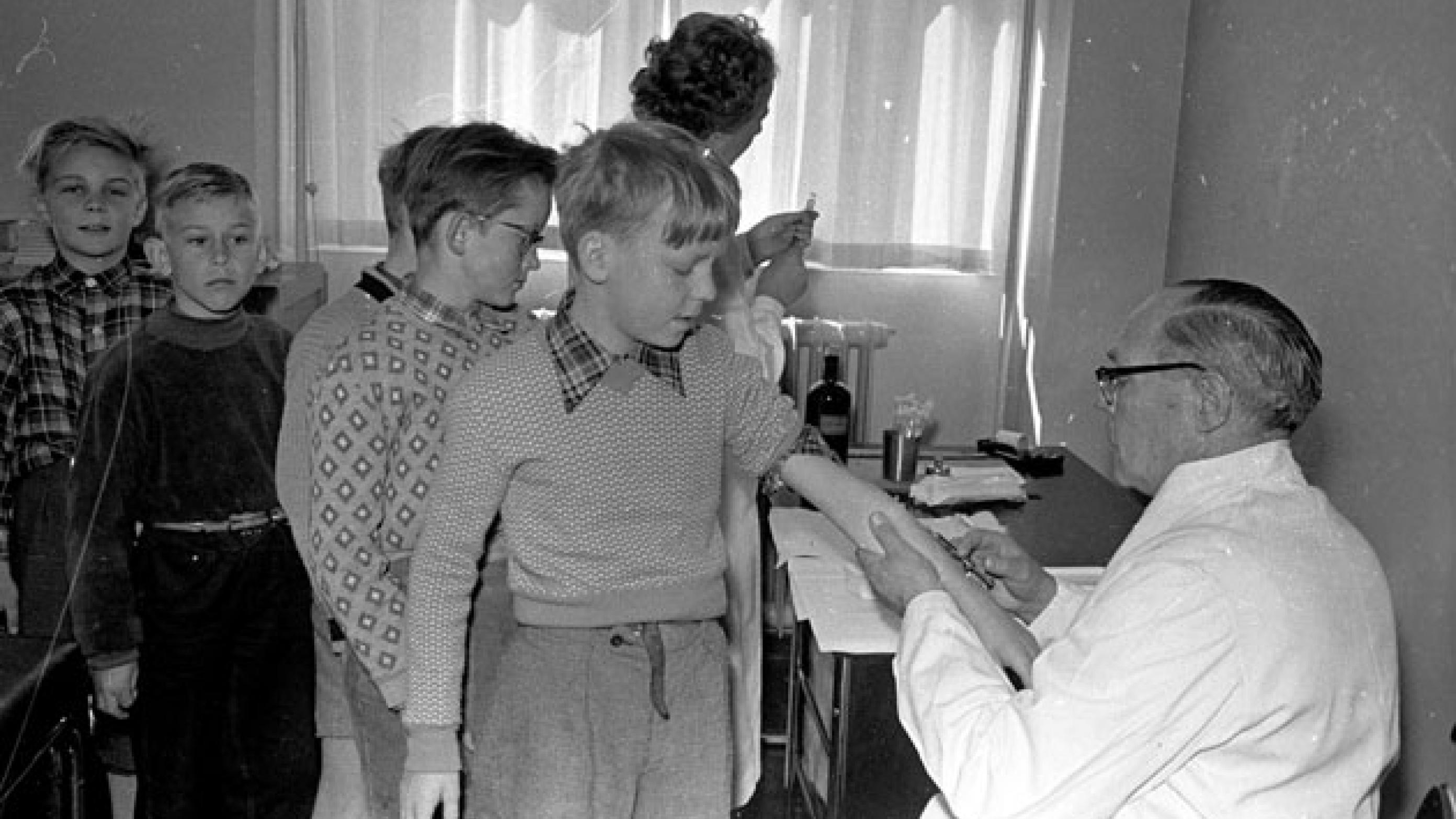 Efter polioepidemien i 1953 var danske forældre panisk angste for, at deres børn skulle blive ofre for en ny epidemi. Her er det amtslæge Hj. Heerup, som i april 1955 stikker nålen i armen på en betænkelig elev på Munkebjergskolen i Odense, mens kammeraterne bagved nervøst venter på deres tur. Foto: Fyens Stiftstidendes pressefotosamling, Odense Stadsarkiv